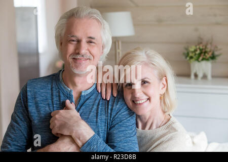 Portrait von Happy senior Paar posieren für Familie Bild zu Hause, Lächeln ältere Frau lehnte sich auf fürsorglichen Mann halten sich an den Händen an Kamera duri suchen Stockfoto