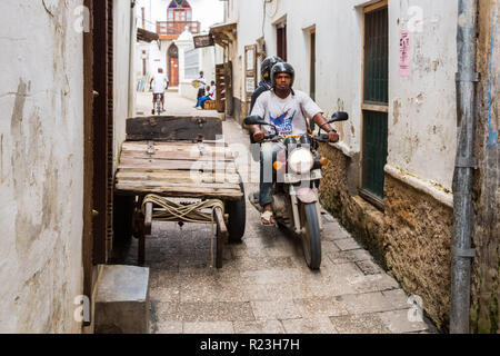 Zwei lokale Männer fahren ein Motorrad durch die engen Straßen von Stone Town, alten kolonialen Zentrum von Zanzibar City, Unguja Insel, Tansania. Stockfoto