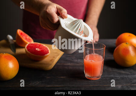 Frisch gepresster Grapefruitsaft ist von einem manuellen Entsafter in ein Glas gegossen. Weißer Kunststoff squeezer in männlicher Hand. Auf einem schwarzen Holztisch sind ganze Ein Stockfoto