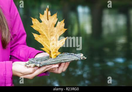 Mädchen in Magenta Jacke holding Holz handgefertigt kleines Spielzeug floss mit eichenlaub als Segel in der Nähe des Flusses auf düsteren Herbst Tag outdoor. Hände closeup Stockfoto