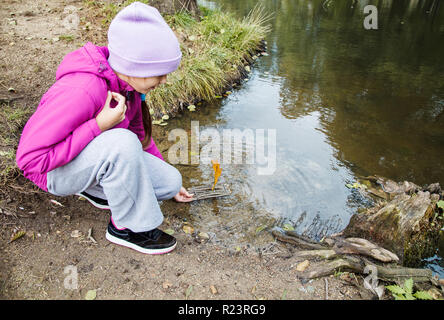 Mädchen im blauen Hut, magenta Jacke und graue Hosen computing Holz handgefertigt kleines Spielzeug floss in den Stream im Freien auf Herbst Tag Stockfoto