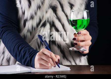 Frau schreiben im Notebook und hält Absinth Alkohol zu trinken in der Hand auf alten retro Holzschreibtisch. Grüne Fee trinken Inspiration für Schriftsteller. Nahaufnahme Stockfoto