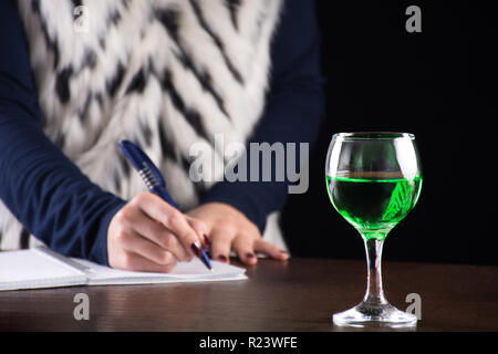 Absinth grüne Getränk in Glas und Frau schreiben Geschichte Notebook im Hintergrund auf alten retro Holzschreibtisch. Grüne Fee Inspiration und Muse für Verfasser Stockfoto