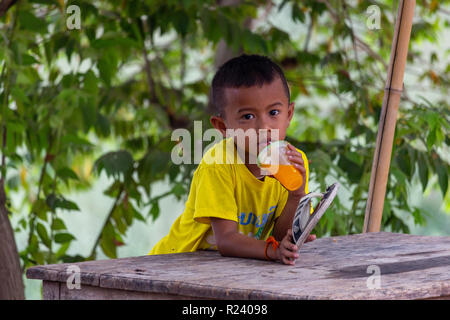 Don Det, Laos - April 24, 2018: Lokale Kind sitzen auf einem Tisch und trinken Orangensaft von grünen Pflanzen und den Fluss Mekong umgeben Stockfoto