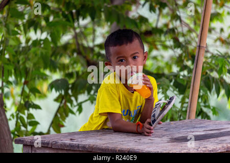 Don Det, Laos - April 24, 2018: Lokale Kind sitzen auf einem Tisch und trinken Orangensaft von grünen Pflanzen und den Fluss Mekong umgeben Stockfoto