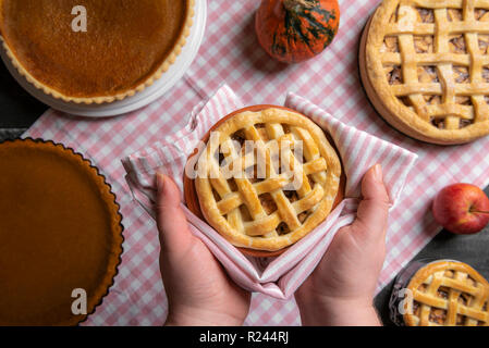 Hände halten ein Fach mit einem Apple Pie, über einen Küchentisch voll von verschiedenen Kuchen, Torten, Kürbis und Apfel Kuchen, auf einem Handtuch. Selbst gebackenem Gebäck. Stockfoto