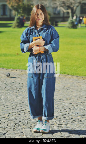 Ein Student in einem denim Anzug steht auf dem Gebiet der Universität und hält ein aufgeschlagenes Buch in den Händen, blickt in die Kamera lächeln Stockfoto