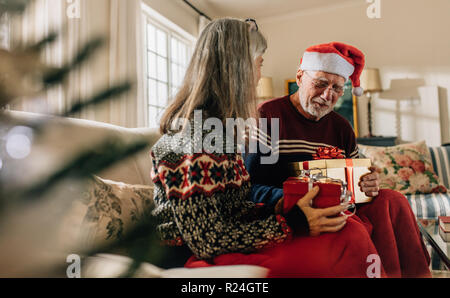 Alte Paar feiert Weihnachten zu Hause und Austausch von präsentiert. Älterer Mann in santa Kappe zu Hause auf der Couch sitzen mit Frau holding Weihnachtsgeschenke. Stockfoto