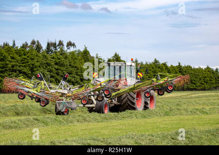 Einen Fendt Traktor und heuwender Arbeiten in einem Bauernhof Feld harken Schnittgras in Zeilen, die für das Heu pressen Stockfoto