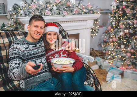 Schönes Bild der jungen Paare zusammen saßen und auf der Kamera. Er hält Fernbedienung. Sie hat Schüssel Popcorn in den Händen. Sie lächeln. Menschen ein Stockfoto