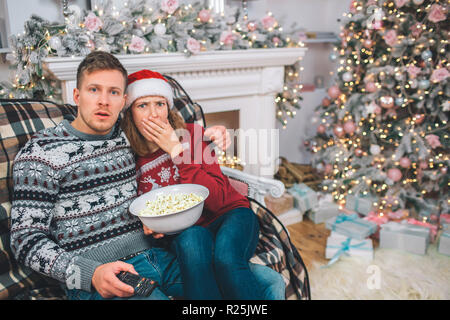 Junges Paar zusammen zu sitzen. Sie sind erschrocken und erstaunt. Junge Frau coveres Mund mit der Hand. Guy ist schockiert. Sie hält Schüssel Popcorn Stockfoto