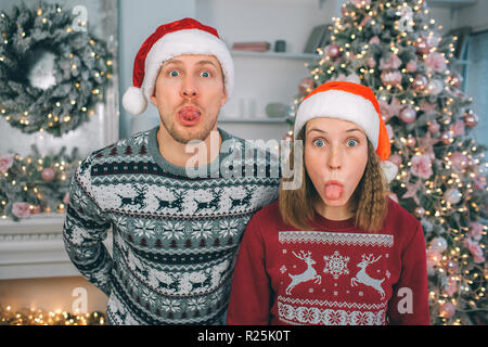Junger Mann und Frau in festlicher Kleidung sehen Sie auf Kamera. Sie stehen zusammen und zeigt ihre Zungen. Kerl hält die Hände hinter sich. Sie tragen Weihnachten Hüte Stockfoto
