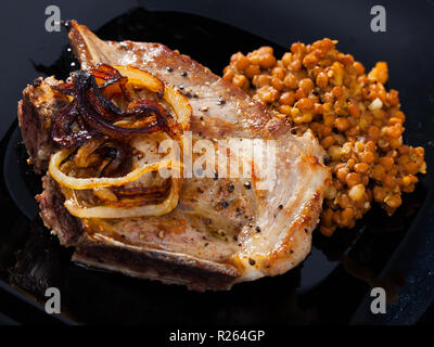 Bild von köstlich gebratenes Schweinefleisch mit Linsen am schwarzen Teller am Tisch Stockfoto