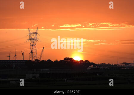 Schönen Sonnenuntergang mit einem orangefarbenen Himmel. Strommast, Stromleitungen und Windenergieanlagen sind gegen den Abendhimmel. Stockfoto