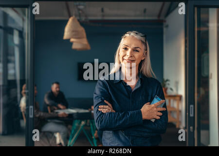 Portrait von lächelnden reife Frau stehend außerhalb Tagungsraum mit Team über die Arbeit im Hintergrund. Lächelnd Senior Business Frau im Büro. Stockfoto
