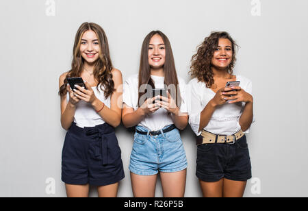 Drei hübsche junge Mädchen legere Kleidung nach oben auf der Suche und Nutzung von Mobiltelefonen über grauer Hintergrund Stockfoto