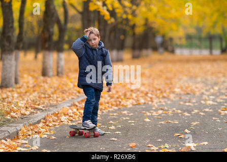 Junge in warme Jacke mit Skateboard seine Hand an die Stirn, als ob sie verletzt ist, allein im Park. Gelbe Blätter und Bäume verschwommenes Ich Stockfoto