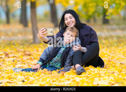 Lächelnde junge Frau und junge Unter einem selfie, als sie zusammen auf die bunten Blätter im Herbst Sit n ein Park mit dem Kind seine Zunge heraus Stockfoto