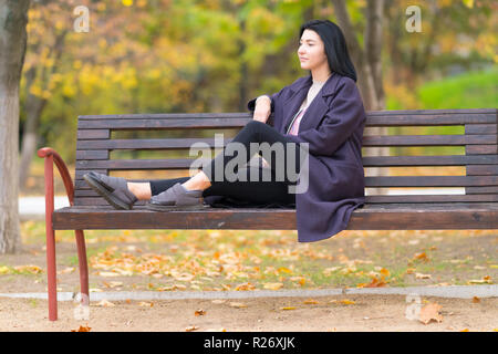 Moderne, junge Frau auf einem rustikalen Holzbank in einem Park nachdenklich an der Seite im Herbst mit bunten Laub hinter starrt Sie Stockfoto