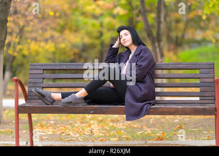 Junge Frau in einem Park denken Tief mit ihren Füßen saß auf einem urigen Holzbank starrte auf die Seite Stockfoto