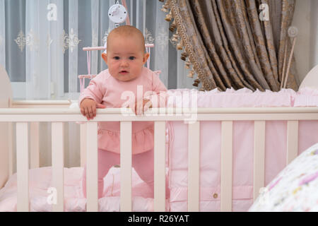 Ein Mädchen stehen und starrte auf die Kamera von einem Gitterbett in ein rosa Schlafzimmer Szene. Stockfoto