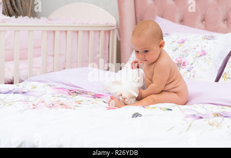 Junge Baby mit ernsten Gesicht ist Spielen mit Spielzeug sitzen auf dem Bett ohne Kleider, Schuß in voller Länge im Zimmer der Eltern mit Kopie Raum Stockfoto