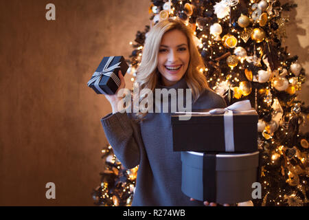 Schöne junge Blondine mit Geschenk unter dem Weihnachtsbaum. Was für das neue Jahr eine junge Frau in einem grauen pullover ihr Geschenke genießt geben Stockfoto