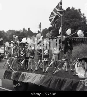 1967, junge Mädchen in Kostümen stand auf dem Rücken eines eingerichteten Schwimmen auf einem Lkw mit Bunting, Ballons und eine Union Jack Flagge bedeckt. Da sie Teil in einem englischen Dorf Straßenkarneval in Oxfordshire, England, UK. Stockfoto