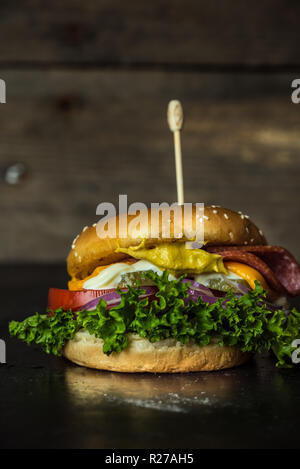 Leckerer cheesburger mit einer großen Anzahl von Gemüse, Senf, Mayonnaise, Salami und lila Zwiebelringe auf Holz, dunklen Hintergrund Stockfoto