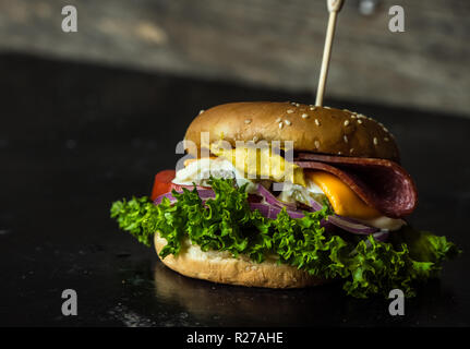 Leckerer cheesburger mit einer großen Anzahl von Gemüse, Senf, Mayonnaise, Salami und lila Zwiebelringe auf Holz, dunklen Hintergrund Stockfoto