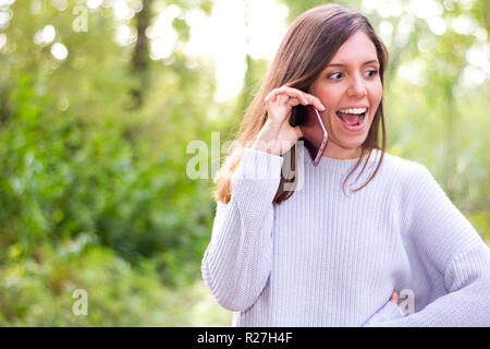 Schöne junge brünette Frau Aufruf per Handy sehr gerne hören zu einem Klatsch in der Natur mit Bäumen Hintergrund Stockfoto