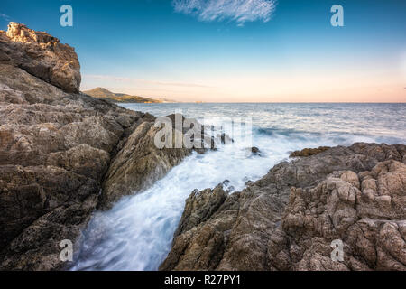 Wellen auf der felsigen Küste von Korsika in lozari als Morgendämmerung bricht über Ile Rousse in der Ferne Stockfoto