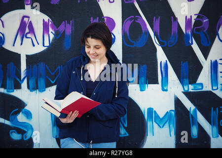 Casual glückliche Frau student Buch lesen, im freien Lebensstil portrait zurück lehnend auf Graffiti Wand Hintergrund. Bildung Konzept, Street Art. Stockfoto