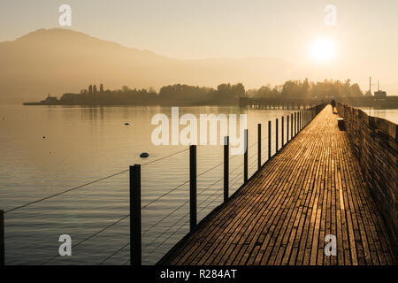 Lange Holzsteg pier über Wasser in goldenen Abendlicht mit einer Berglandschaft und Menschen zu Fuß Silhouette im Hintergrund Stockfoto