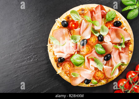 Lecker lecker Pizza mit Käse, Tomaten, Rucola, Oliven, Basilikum und italienischen Schinken auf dunklem Stein Hintergrund. Blick von oben. Mit Horizontal Stockfoto