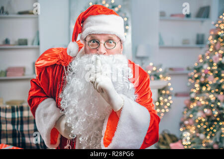 Erstaunt Santa Claus hält mit der einen Hand frint von weißen Bart und sieht auf der Kamera, obwohl eine Brille. Er hat rote Tasche über der Schulter. Stockfoto