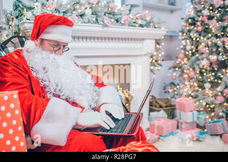 Moderne Santa Claus sitzt am Kamin und Weihnachtsbaum. Er hält das Laptop ein nap und funktioniert. Guy ist konzentriert. Stockfoto