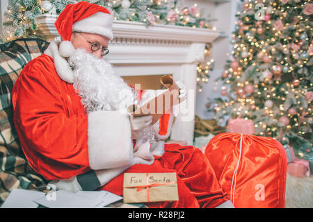 Ernster und nachdenklicher Mann in Weihnachten suite sitzt und liest Briefe. Es gibt andere neben ihm auf dem Tisch. Man sitzt am Kamin und Weihnachtsbaum. Stockfoto