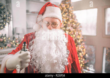 Entspannt Mann in Santa Claus Kostüm steht und hält vape in der Hand. Tritt Rauch aus seinem Mund. Er ist entspannt. Stockfoto