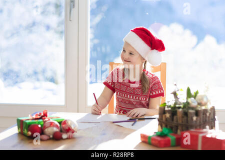 Kind schreiben Brief an Santa am Heiligabend. Kinder schreiben Weihnachtsgeschenk Wunschliste. Kleines Mädchen sitzen in eingerichtetes Wohnzimmer mit grossen Fenster auf Sun Stockfoto