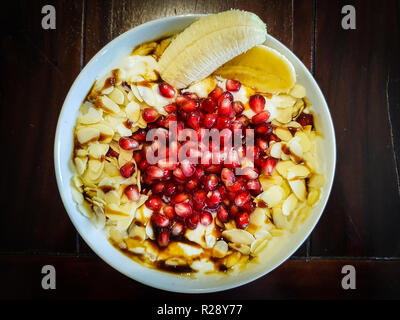 Eine Schüssel voll mit Müsli mit Hafer Früchte Banane Granatapfel Mandeln Joghurt und Ahornsirup Stockfoto