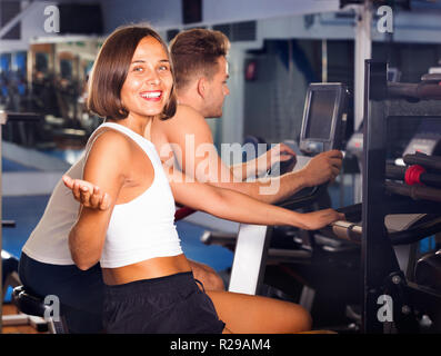 Positive junge Frau und mit Fahrradverleih Fitnessraum Maschinen gemeinsam drinnen Mann Stockfoto