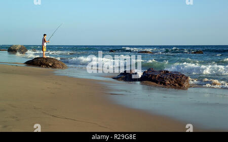 Ein junger Mann Fischer stehen auf einer Küstenlinie rock versucht sein Glück bei Sonnenuntergang als Pacific Ocean Surf Rollen auf dem sandigen Strand an der Crystal Cove State Park in Newport Beach, Kalifornien, USA. Stockfoto