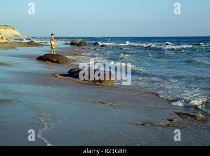 Ein junger Mann Fischer stehen auf einer Küstenlinie rock versucht sein Glück bei Sonnenuntergang als Pacific Ocean Surf Rollen auf dem sandigen Strand an der Crystal Cove State Park in Newport Beach, Kalifornien, USA. Stockfoto