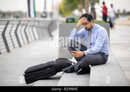 Arabische Geschäftsmann mit seinem Koffer in einer Stadt, sitzt auf der Treppe und suchen bei seinem Telefon Stockfoto