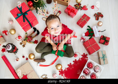 Neugierige Mädchen mit Weihnachten kostüm Rentier Geweih auf dem Boden sitzend, Weihnachtsgeschenk, Ansicht von oben. Stockfoto