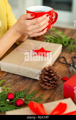 Nicht erkennbare Frau sitzt an einem Tisch mit schön verpackt Weihnachtsgeschenk, holding Tasse Tee. Geschenkpapier xmas-Konzept. Stockfoto