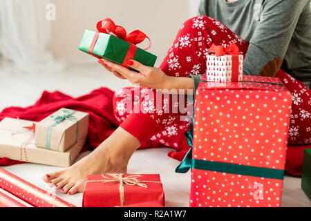 Junge Frau mit xmas Schlafanzug auf dem Boden sitzend unter aufgewickelte Weihnachtsgeschenke, entspannend. Stockfoto