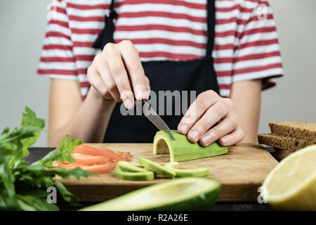 Schneiden Avocado für gesunde Sandwich. Frau Hände Schneiden reif avocado Frucht am Küchentisch mit frischen Zutaten Stockfoto