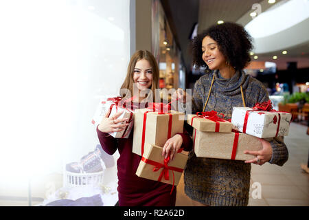 Zwei gemischten Rennen Frauen mit geschenkboxen in den Händen in der Nähe von storewindow. Multi-ethnische Mädchen lächelnd mit Geschenken zu Weihnachten neues Jahr verkaufen. Kaukasische und afrikanische amerikanische Frauen kaufen Geschenke für die Feiertage. Stockfoto